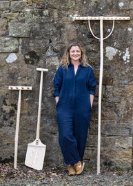 A woman in overalls is smiling at the camera, leaning against a stone wall. She is standing next to a shovel and other gardening tools.