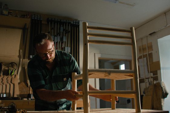 A furniture maker is in their workshop, looking at a small Ash chair on the table.