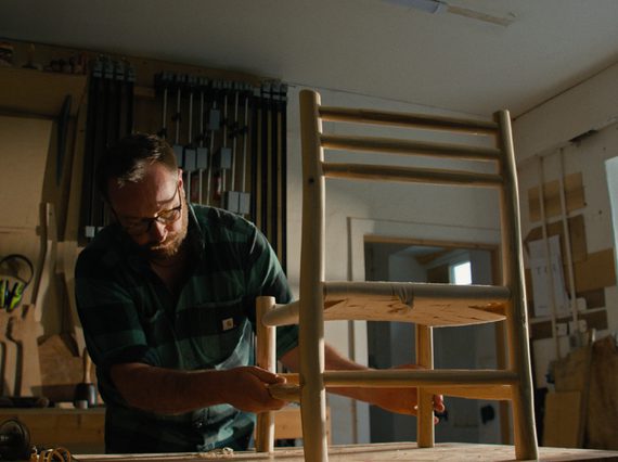 A furniture maker is in their workshop, looking at a small Ash chair on the table.