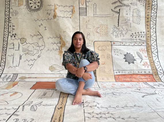 A woman sits holding her legs crossed on a large piece of cloth which extends up behind her. It is covered in hand printed or painted illustrations of leaved and plants.