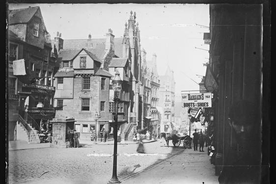 Black and white image of a cobblestone Victorian high street. There are various shop fronts, a horse and cart, and people walking around.