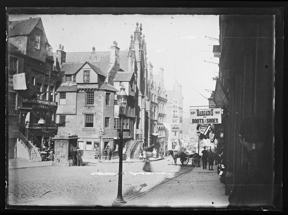 Black and white image of a cobblestone Victorian high street. There are various shop fronts, a horse and cart, and people walking around.