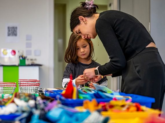 A young person and their carer are standing in a room, at a table covered in crafting materials. They are both looking at something in the older person's hands.