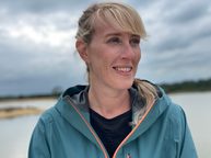 A woman with blonde hair is wearing a blue raincoat, and looking off to the right of the camera. It looks like she's standing on a beach, or near a body of water.