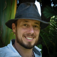 A man, wearing a trilby hat, is smiling at the camera and standing in front of a background of leaves