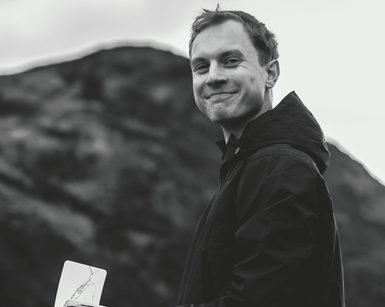 A black and white photograph of a smiling man wearing a waterproog jacket and holding a sketchbook.