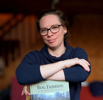 A woman wearing a blue long-sleeve shirt and glasses. They have their arms cross and are leaning on a book titled 'Bog Fashion'.
