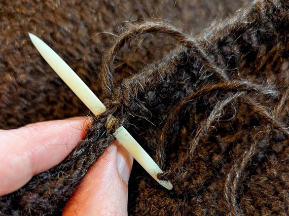 A hand holding a needle that has been threaded through brown wool fabric