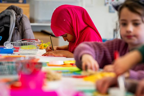 A child drawing at a table with other children doing crafts.