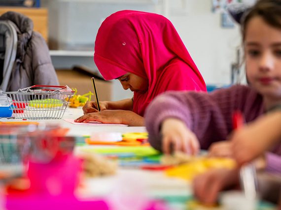 A child drawing at a table with other children doing crafts.