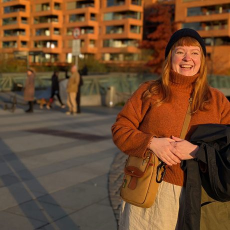 Photograph of Hannah Kelly. She is standing in front of a block of flats. She has long, red hair, and is wearing a black beanie hat with an orange jumper and cream trousers.