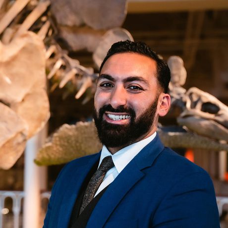 A close up of a man with a beard, in a suit is smiling in front of a dinosaur skeleton on display.