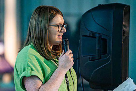 A person standing on a stage with a speaker in the back, holding a microphone.