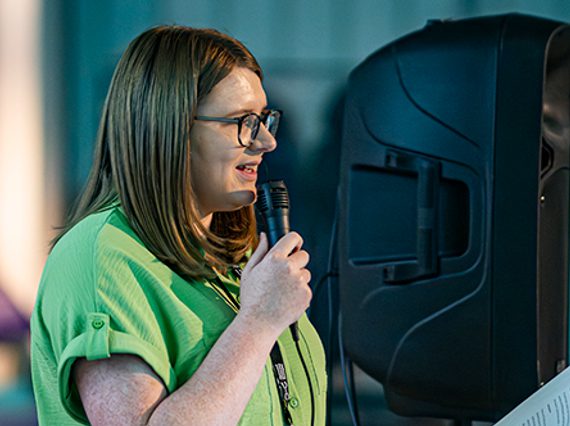 A person standing on a stage with a speaker in the back, holding a microphone.