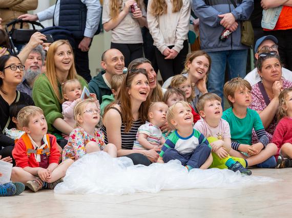 A group of children sit with their carers and parents on the floor at the museum. They're gazing up at something being performed behind the camera.
