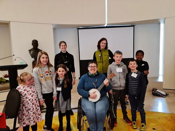 A group of children stand around a person sitting in a wheelchair playing a banjo