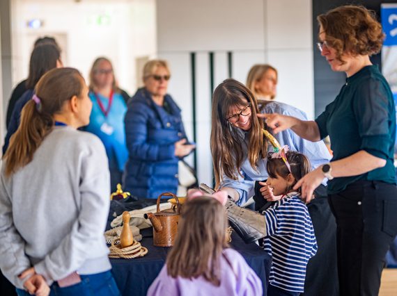 Children looking at objects like a watering can and a knight's armored gloves on a table, surrounded by adult helpers.