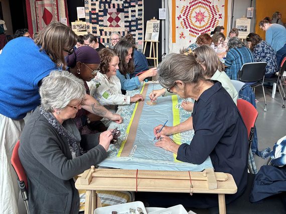 A group of people sit at a table quilting fabric.