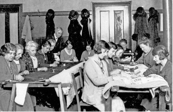 A black and white photograph of a group of women sit around long tables quilting.
