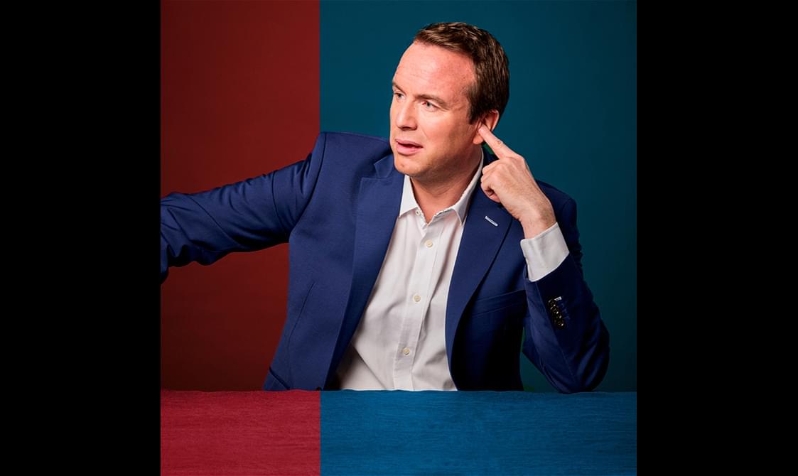 Matt Forde sits in front of a blue and red background wearing a blue suit.