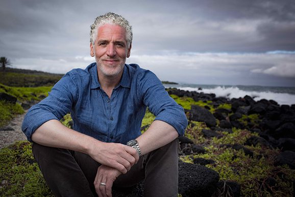 A man sits cross legged, looking into the camera. In the background there's a cloudy sky, and green vegetation.