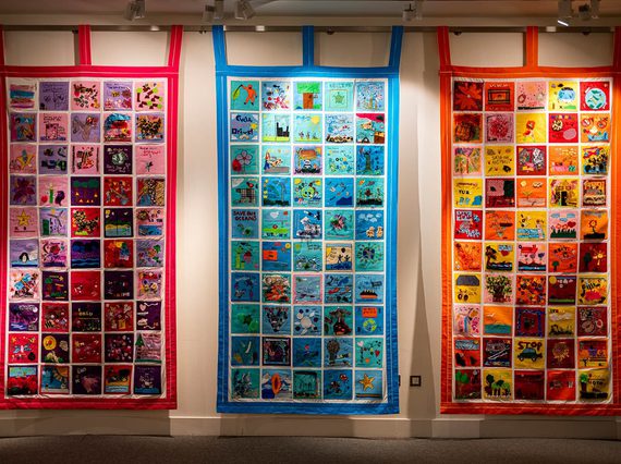 Three large vibrant quilts hanging against a yellow wall. Made up of colourful squares showing patterns, shapes and words of hope for the future.