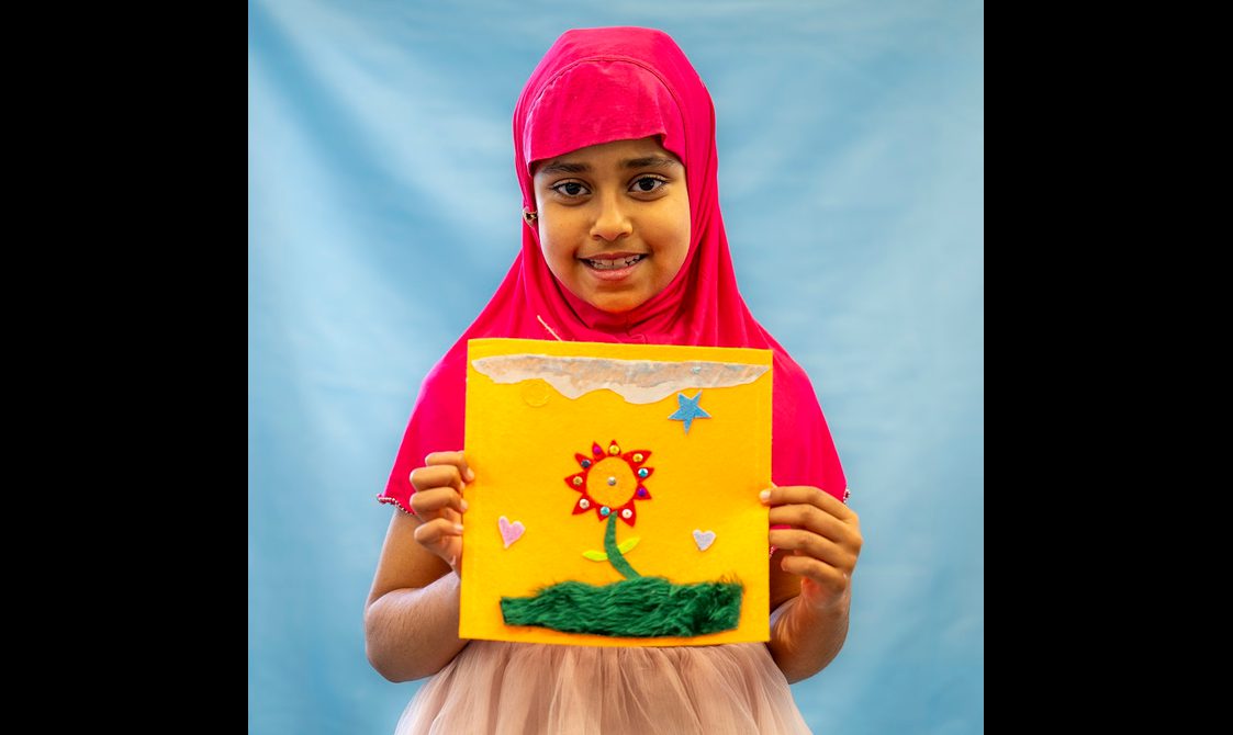A young person is smiling directly at the camera, holding up a quilt square with an image of a flower.
