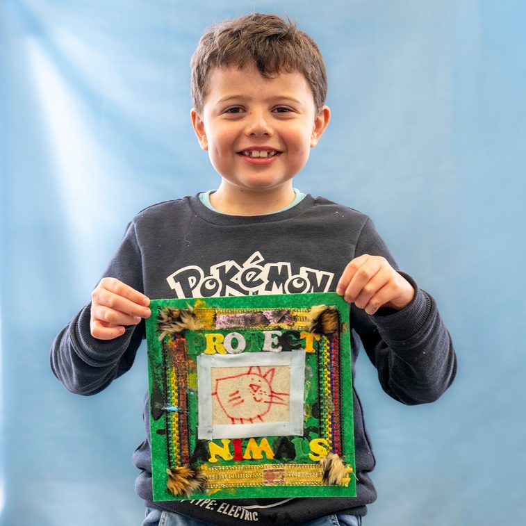 A young person is smiling directly at the camera, holding up a fabric quilt scare that reads 'protect the animals'.