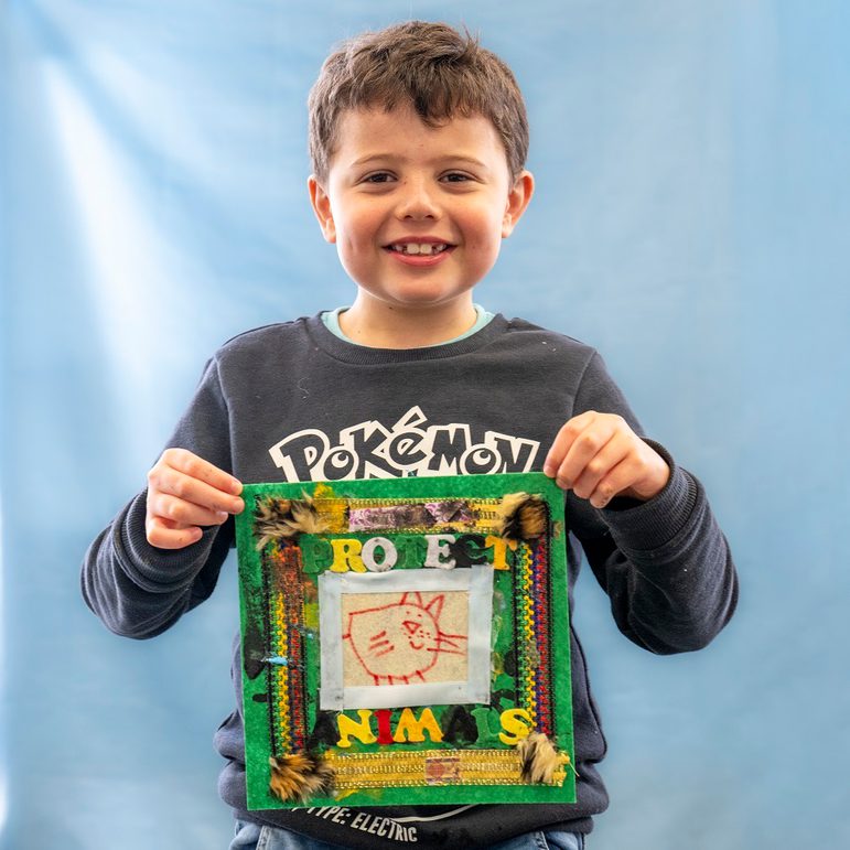 A young person is smiling directly at the camera, holding up a fabric quilt scare that reads 'protect the animals'.