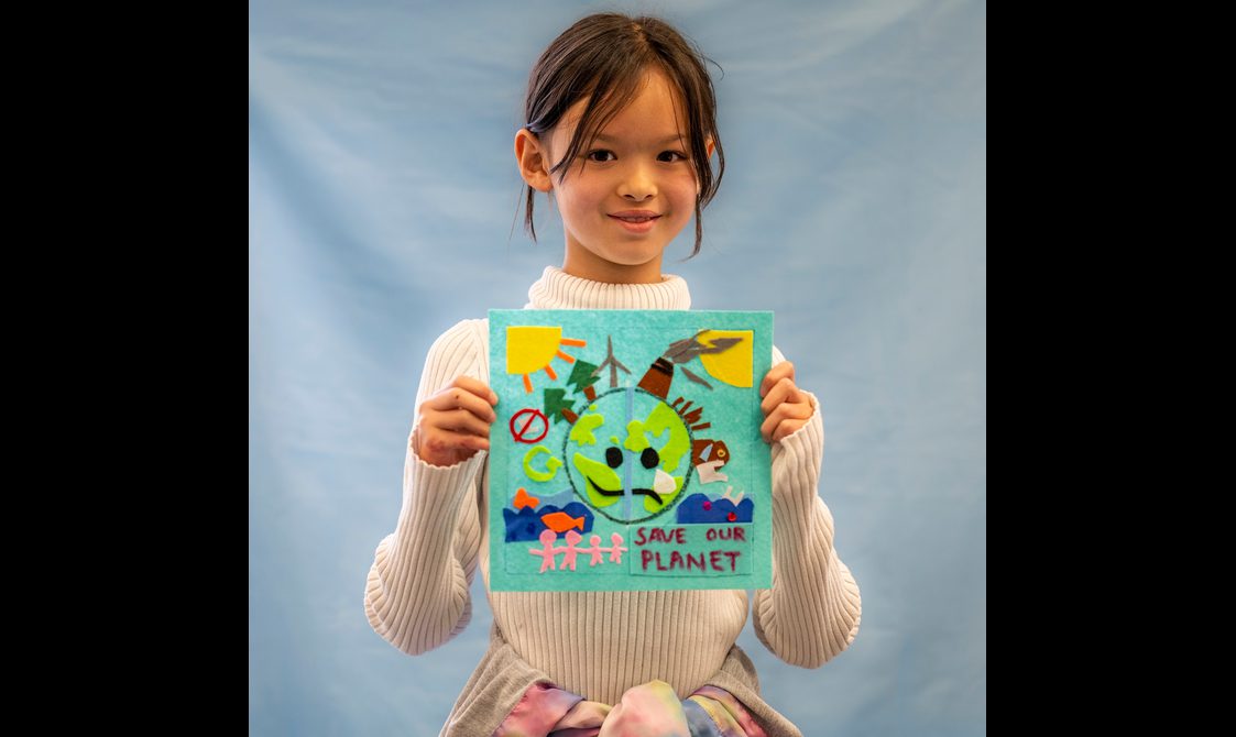 A young person is holding a quilt square up to the camera, and smiling.