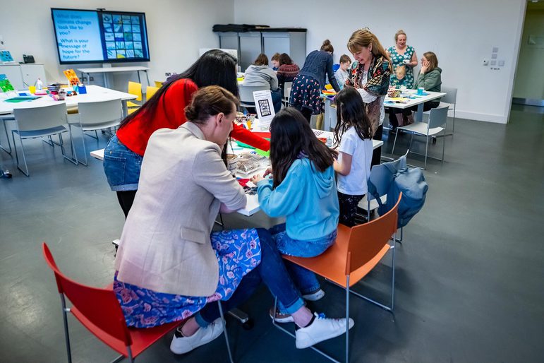 Group of adults and children sitting at tables doing quilt crafts.