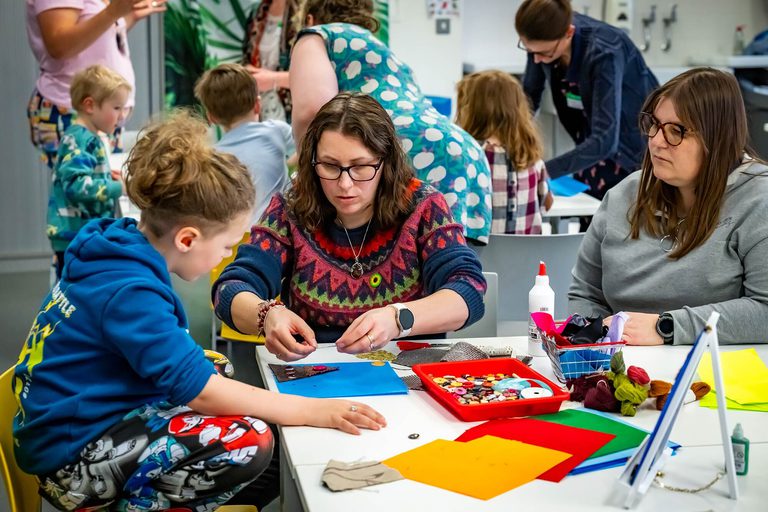 Two adults and a child sit at a table doing a quilting craft activity.