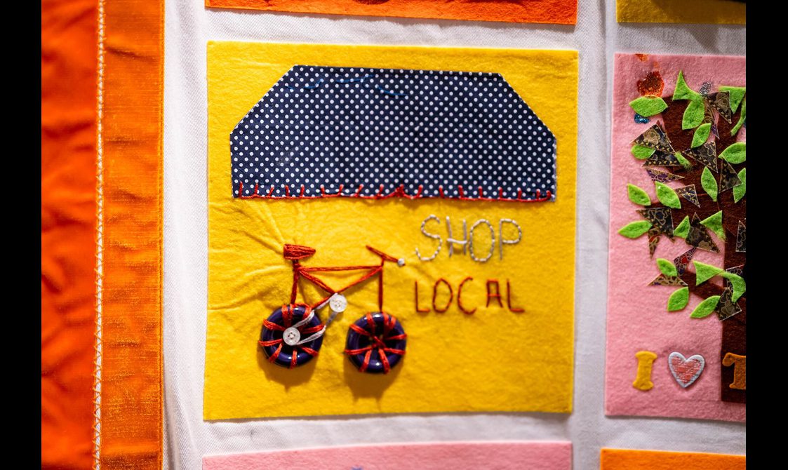A yellow quilt square with the message "shop local" sewn on it. There is a small red bike sewn into the fabric, with blue buttons for wheels.
