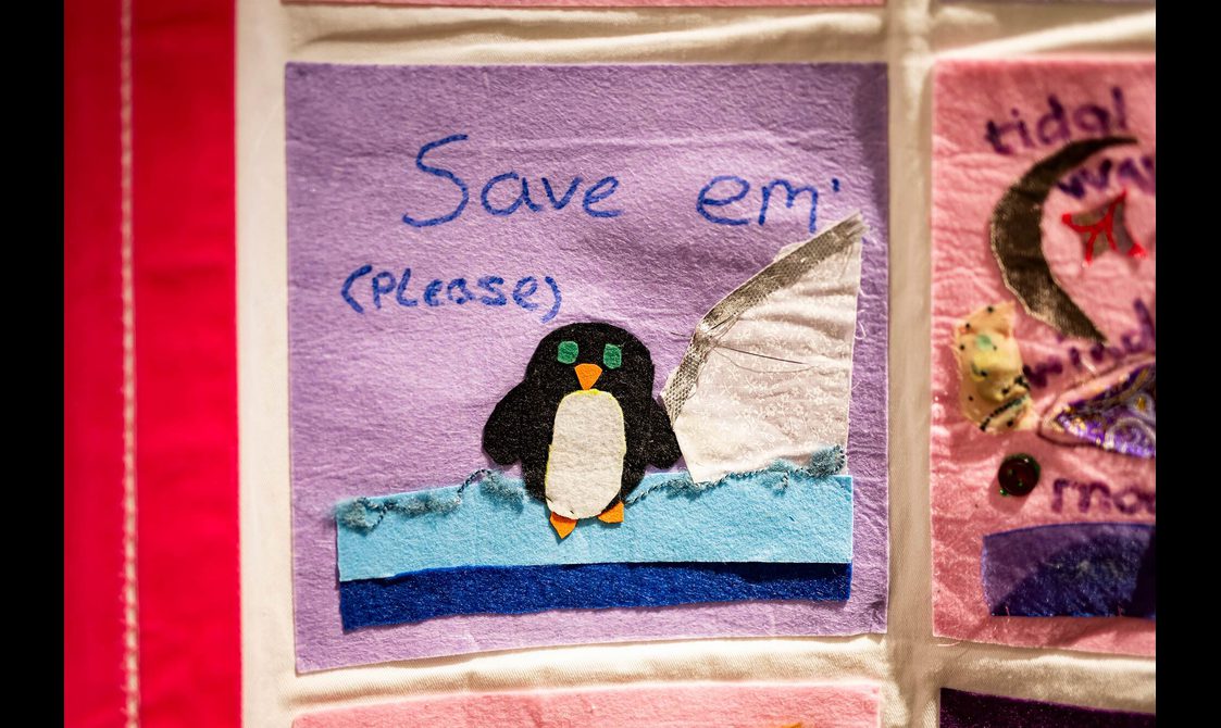 A purple quilt square with a small felt penguin standing on a snowy landscape. The message "Save 'em please" is written above the penguin.