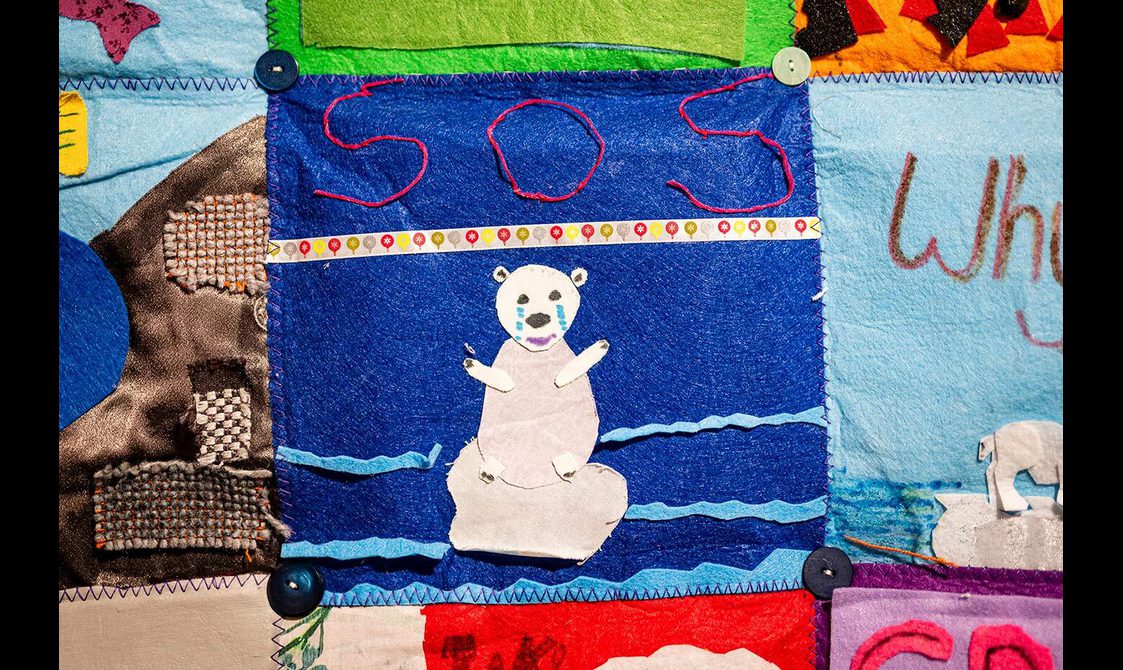 A blue quilt square with a polar bear made from different pieces of fabric. The polar bear is standing on a melted ice cap, crying. Above the polar bear reads the message "SOS" in read letters.