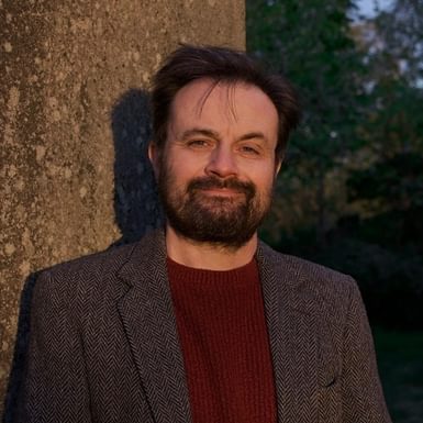 A man is wearing a brown blazer and wine red tshirt, and is standing with his back to a tree and smiling at the camera.