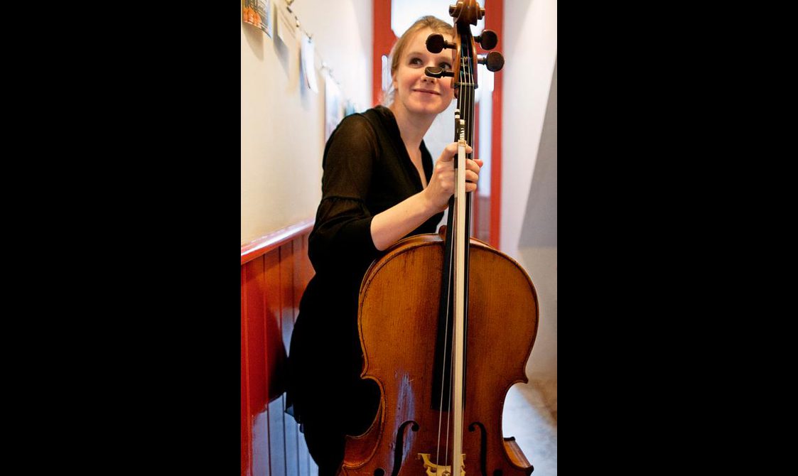 A woman holding a cello in a corridor, with the top of the cello partially obscuring her face.