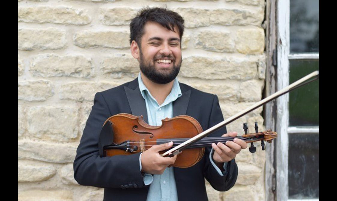 A man in front of a brick wall, smiling and holding a fiddle.