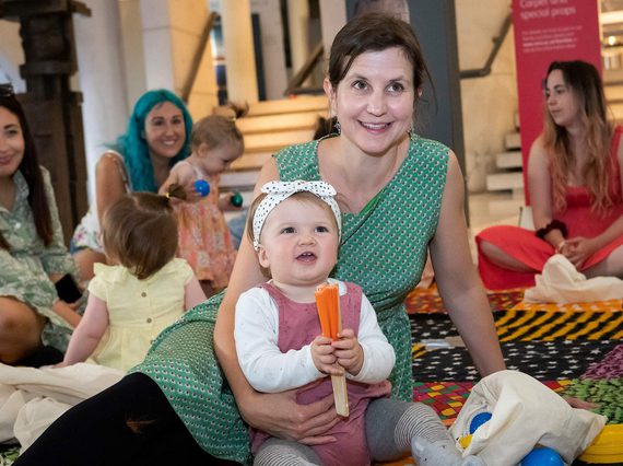 A parent and toddler sitting on a colourful carpet playing with toys. Other parents and toddlers are sitting in the background.