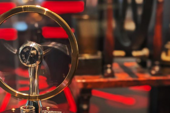 A brass and black small replica of a Stirling Engine is held up in front of a case. The background is an out of focus room filled with red and black stripes.