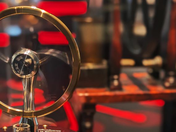 A brass and black small replica of a Stirling Engine is held up in front of a case. The background is an out of focus room filled with red and black stripes.