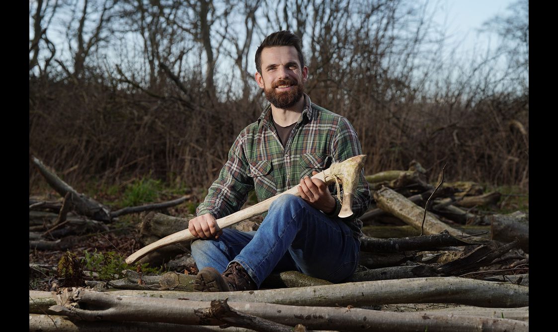 A man with dark hair and a bear wears a green tartan shirt and blue jeans. He sits on a pile of logs, and holds a replica of a Bronze Age axe.