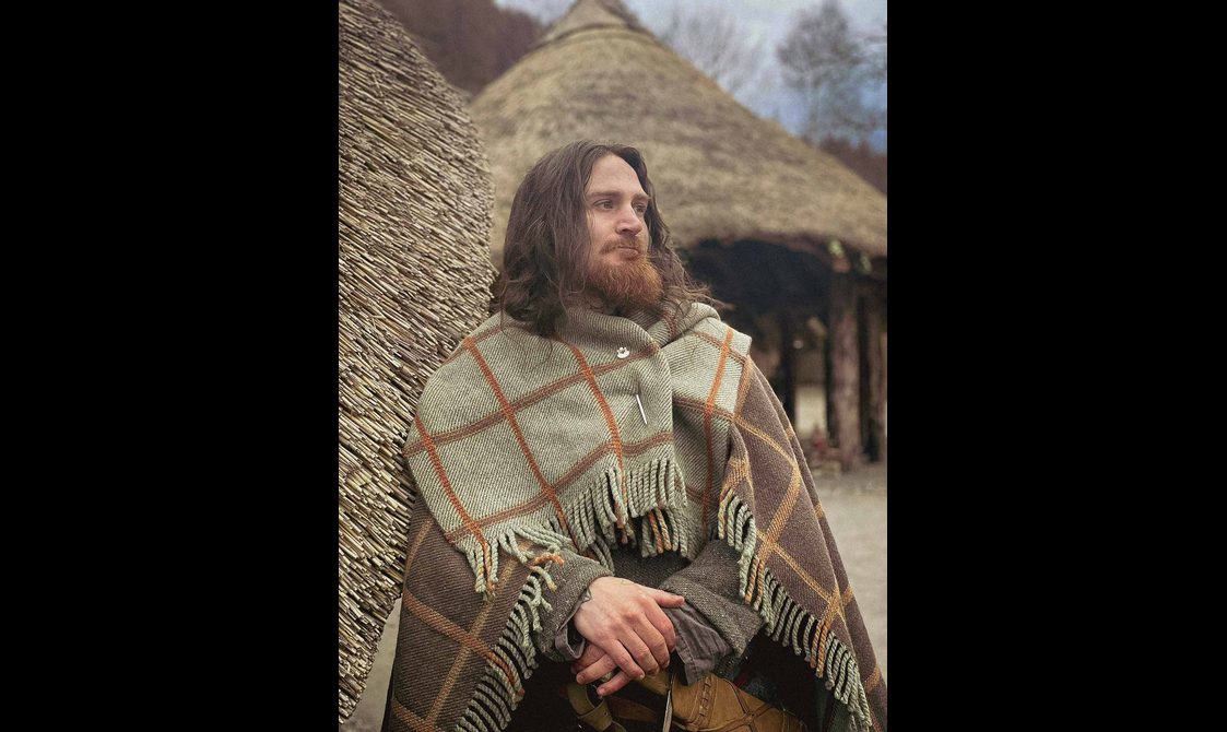 A man with long brown hair and a beard wears a green and brown tartan cloak.