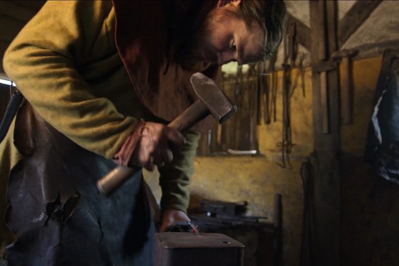 A blacksmith wearing a beige apron hits metal with a hammer.