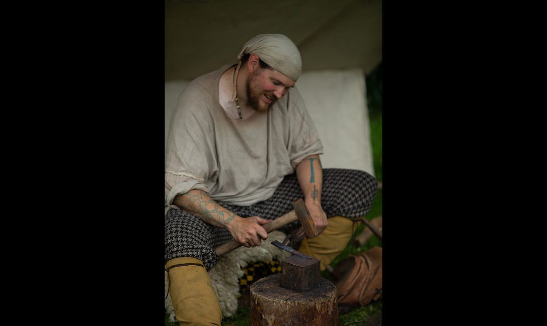 A man wearing a grey shirt work with a hammer and metal.