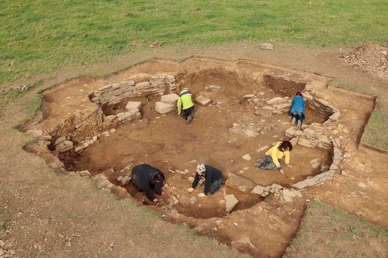 Aerial view of large square of brown dirt in a green field dug out to reveal archaeological burial site, with people excavating.