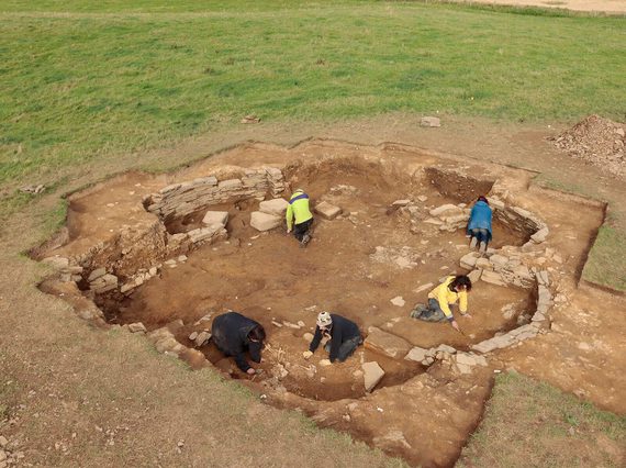Aerial view of large square of brown dirt in a green field dug out to reveal archaeological burial site, with people excavating.