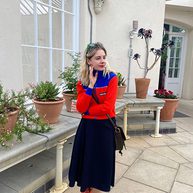 A woman with long blonde hair, a red top, and black skirt poses in front of plotted plants.