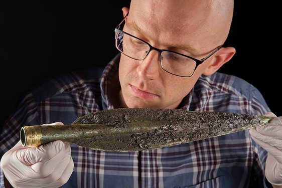 A man in a shirt, glasses and white gloves examines a corroded bronze age spear head in front of a black background.