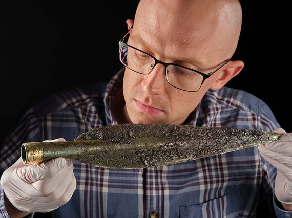 A man in a shirt, glasses and white gloves examines a corroded bronze age spear head in front of a black background.